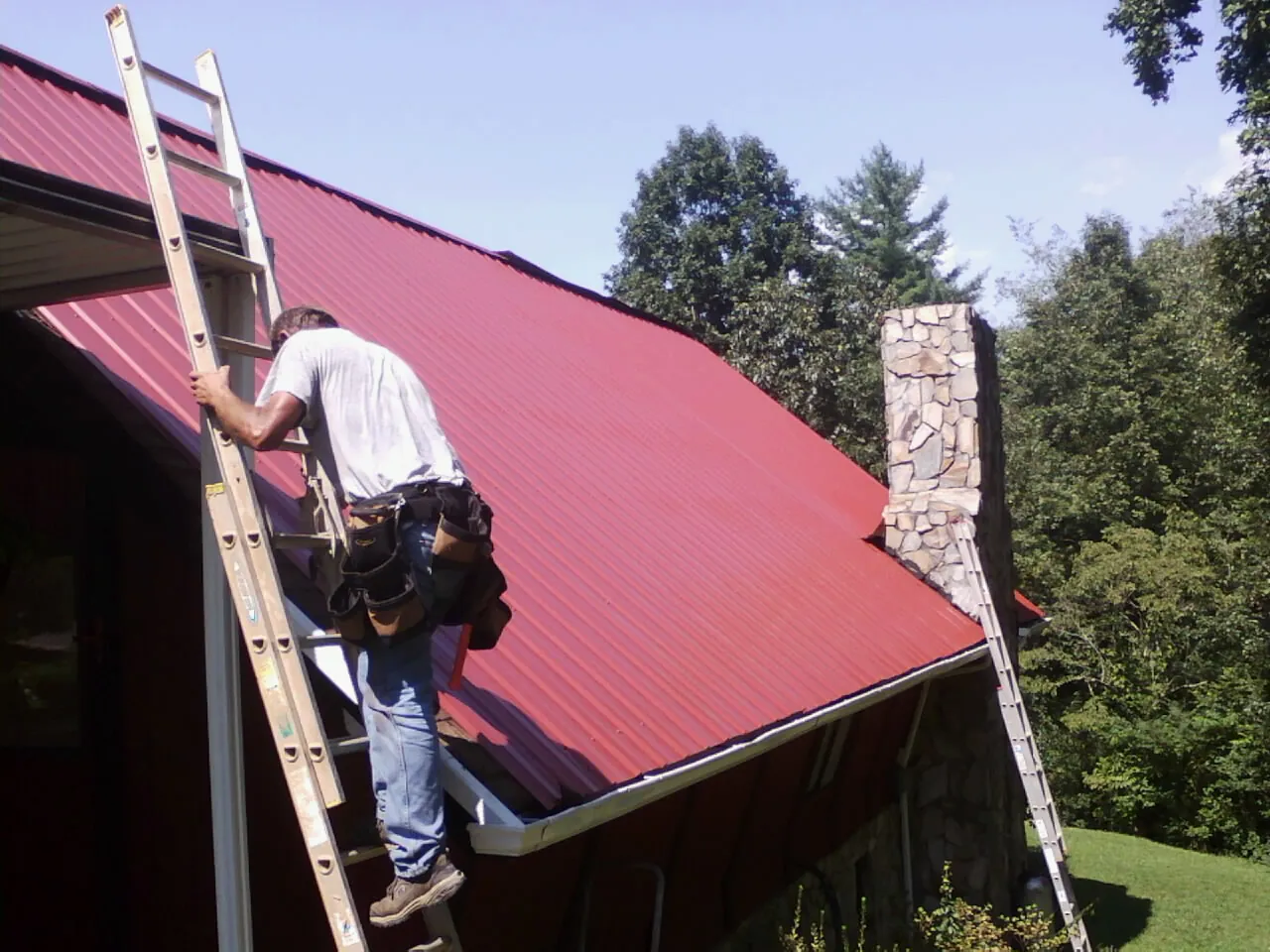 Roofer on a ladder beside a red metal roof and stone chimney — Soffit & Fascia Repair by Roofing Brothers in Imbler